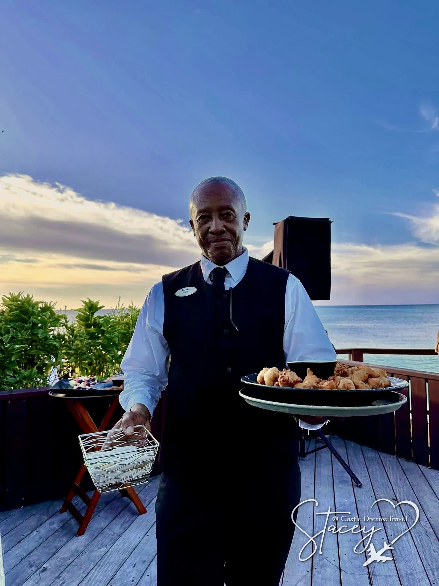 A Beaches server presenting a tray of appetizers on an oceanfront deck at sunset