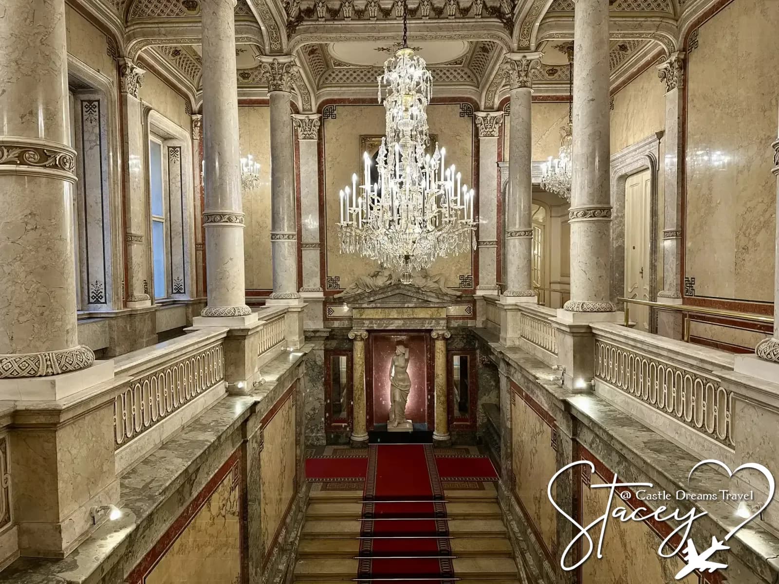 An ornate marble staircase in Vienna beneath a crystal chandelier, with a red carpet runner