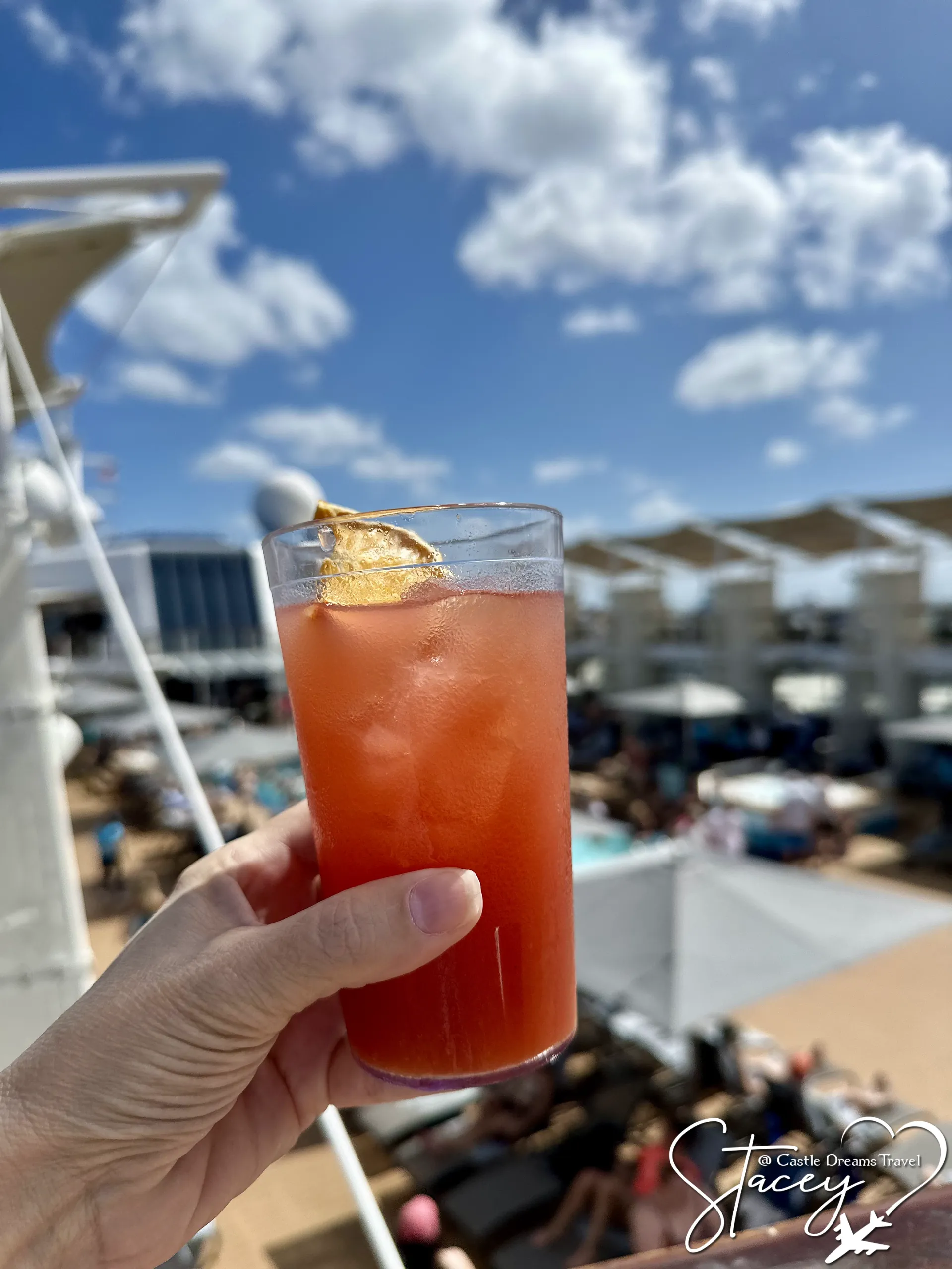 A tall iced cocktail held up on a Celebrity Cruises pool deck under blue sky and scattered cloud