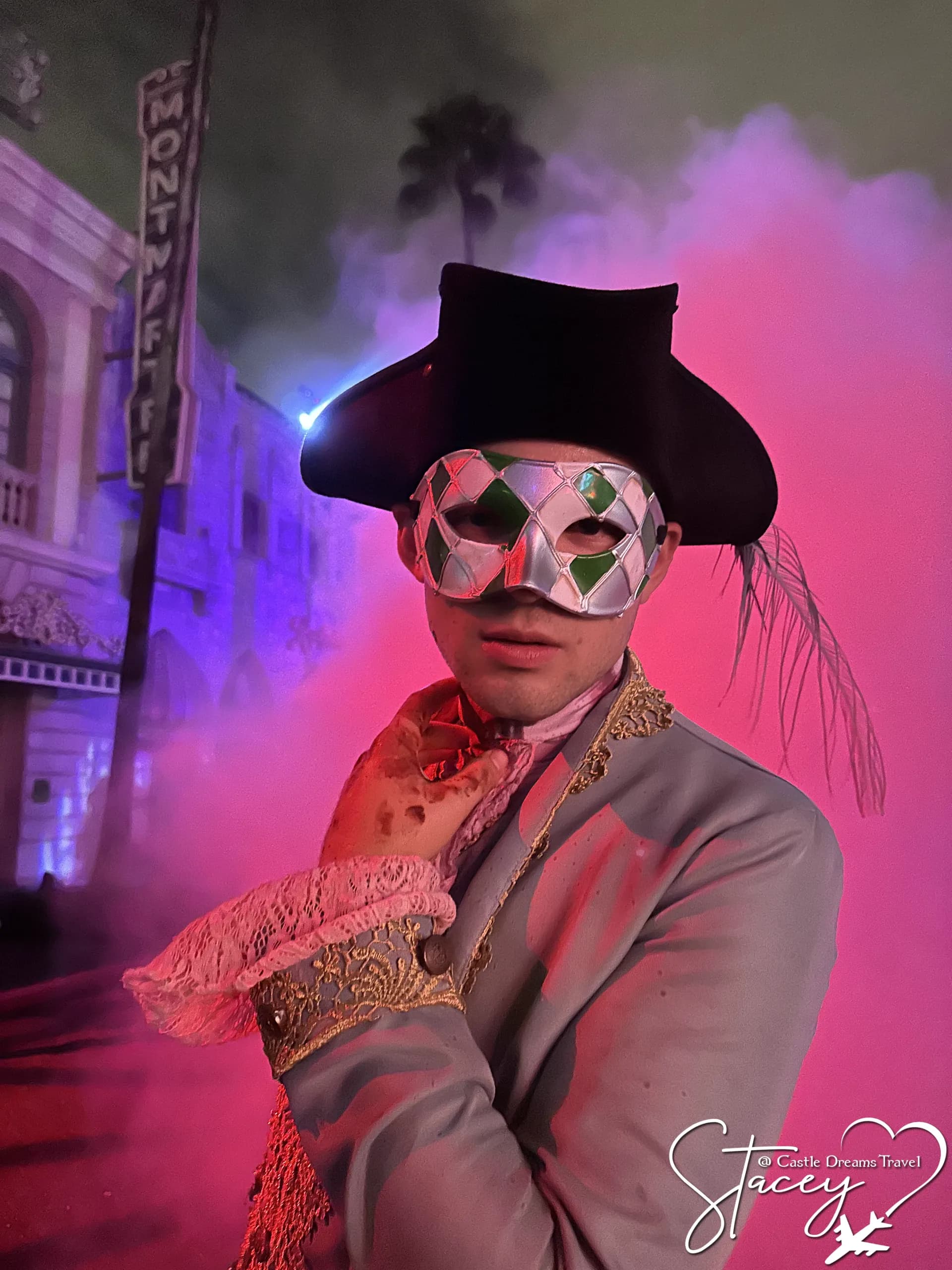 A performer in a plumed tricorn hat and harlequin mask standing in pink fog