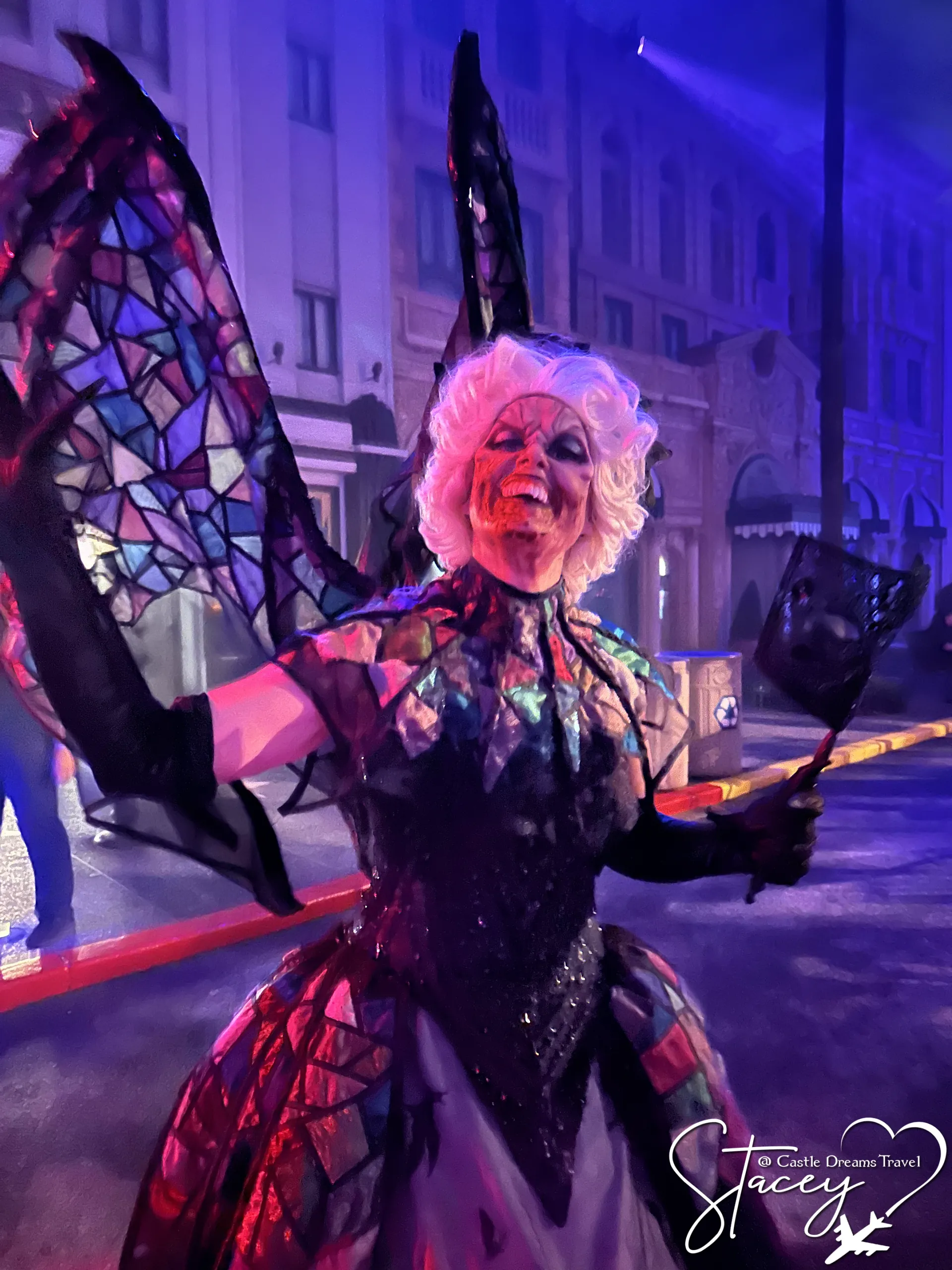 A performer in a stained-glass winged costume and pale wig posing under blue light