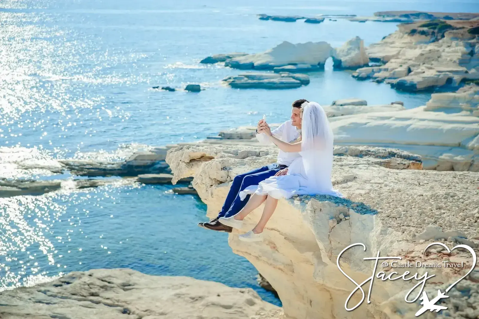 A bride and groom sitting together on white coastal rocks above a sparkling blue sea