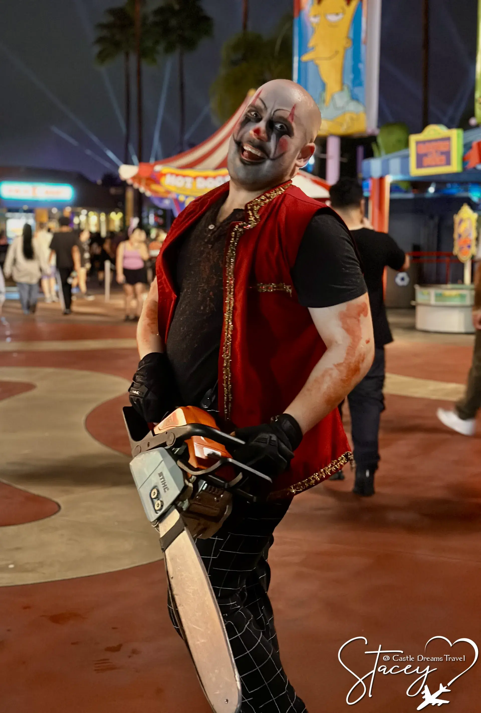 A scare actor in clown makeup and a red vest carrying a chainsaw along a Halloween Horror Nights street