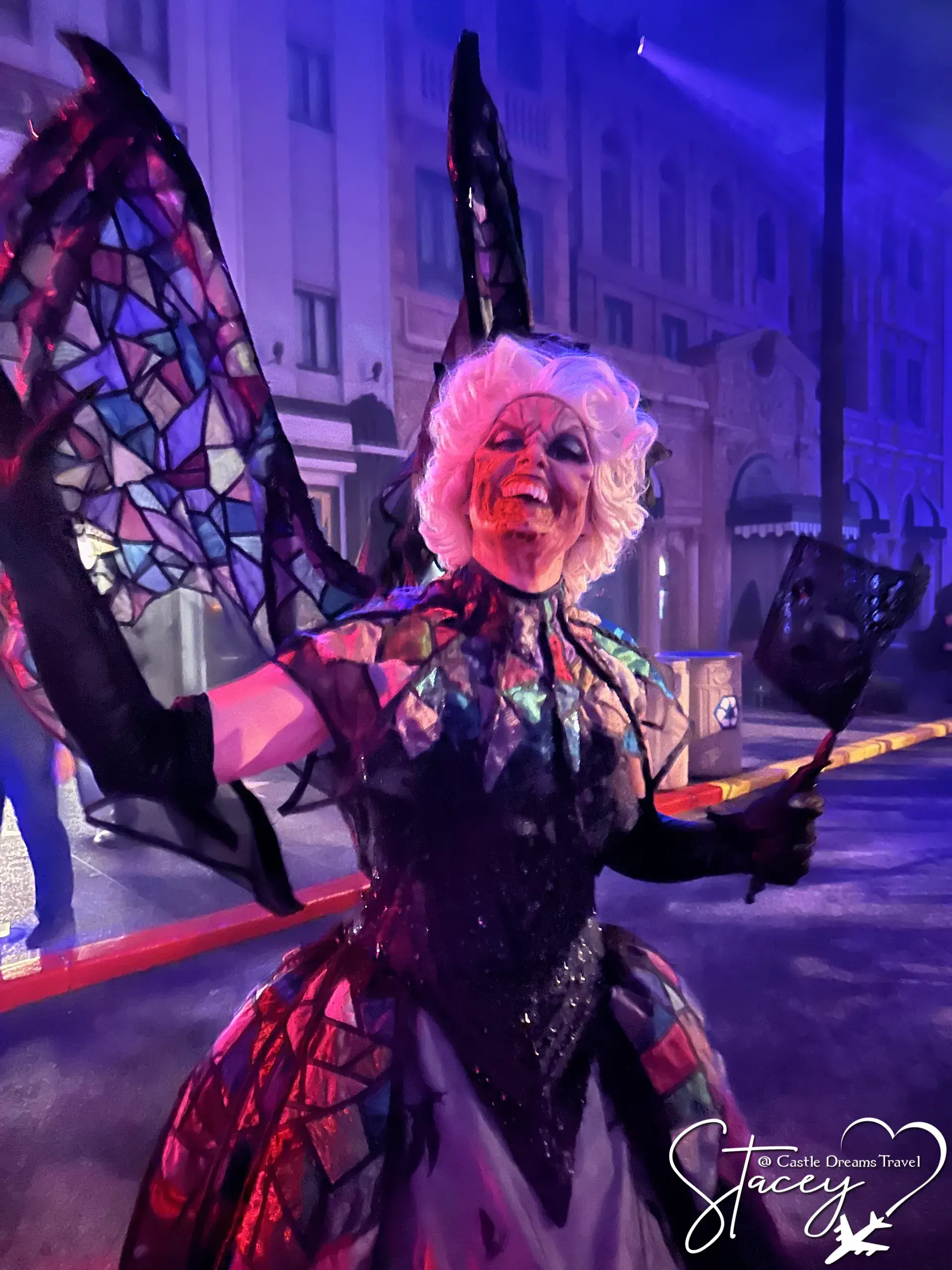 A performer in a stained-glass winged costume and pale wig posing under blue light